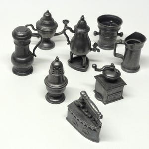 May include: A collection of eight miniature pewter-colored metal household items including a coffee pot, a tea kettle, a pitcher, a salt shaker, a coffee grinder, and a flat iron. Each piece features a dark grey metallic finish and detailed molding.