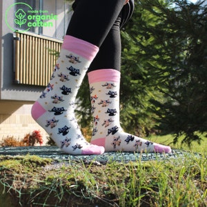 Op de afbeelding: Crèmekleurige sokken met een herhaald patroon van cartoonkatten in verschillende kleuren. De sokken hebben roze hielen en manchetten. Een groen logo linksboven toont "made from organic cotton".