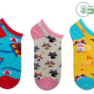 May include: Three pairs of no-show socks made from organic cotton. One pair features a beach scene, another has a pattern of cats, and the third has a cat wearing sunglasses. The socks have red, pink, blue, and yellow accents.