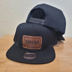 Pode incluir: Duas bonés de beisebol pretos, um com um remendo de couro marrom que diz "BANGTAN" e "VII.XII.MMXIII". O boné de baixo tem uma etiqueta preta "SNAPBACK".