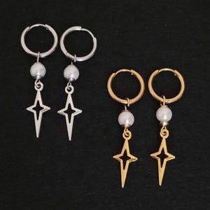 May include: Two pairs of earrings on a black background. One pair is silver-toned with a pearl bead and a star pendant. The other pair is gold-toned with a pearl bead and a star pendant. Both pairs have hoop closures.