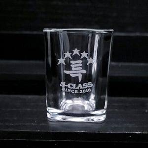 May include: A clear glass shot glass with a square base. The glass has an etched design of five stars, a stylized character, and the text "S-CLASS SINCE 2018".