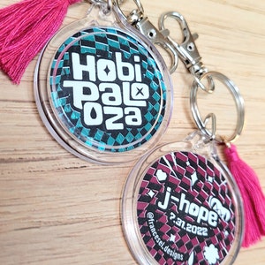 Op de afbeelding: Twee ronde sleutelhangers met doorzichtige kunststof frames en kleurrijke ontwerpen. De eerste sleutelhanger heeft een teal en zwart geruit ontwerp met de tekst "Hobi Palooza". De tweede sleutelhanger heeft een rood en zwart geruit ontwerp met de tekst "j-hope 2.31.2022" en "@francesel.designs".