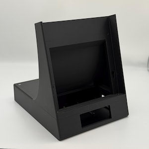 Puede incluir: Una carcasa metálica negra, rectangular, con una sección superior en ángulo. La carcasa tiene una abertura rectangular en la parte delantera y otra más grande en la parte superior. La base es plana y rectangular.