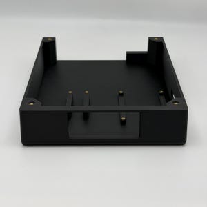 以下が含まれることがあります： 真鍮色のネジインサートが付いた黒色の長方形の電子部品エンクロージャ。エンクロージャの片側にはくぼんだ領域があり、電子部品を収容するように設計されています。全体的なデザインはミニマリストで機能的です。