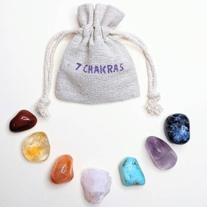 Peut inclure: Sept pierres roulées de chakra dans un petit sac en toile blanc avec le texte "7 CHAKRAS" brodé sur le devant. Les pierres sont disposées en cercle sur un fond blanc. Les pierres sont rouge, orange, jaune, verte, bleue, indigo et violette.
