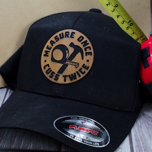 May include: Black baseball cap with a brown leather patch that reads "Measure Once, Cuss Twice". The patch features a hammer and a tape measure. The cap has a Flexfit label and is size L/XL.