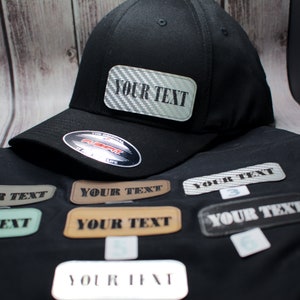 Op de afbeelding: Zwarte baseballpet met een rechthoekige patch met de tekst "YOUR TEXT" in het wit. Verschillende patches met dezelfde tekst worden onder de pet weergegeven. De patches hebben verschillende kleuren en texturen.