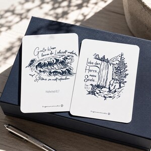 Könnte beinhalten: Zwei weiße Karten mit dunkelblauen Illustrationen und Text. Eine Karte zeigt Wellen mit Text, die andere einen Wasserfall mit Text. Die Karten liegen auf einer dunkelblauen Box mit einem silbernen Stift.