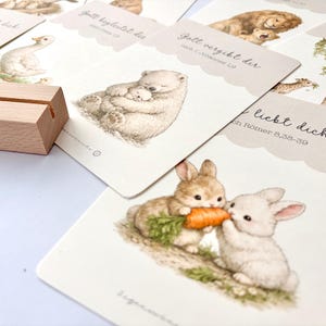 May include: A collection of illustrated cards featuring various animals, including a bear with a cub, a lion with a cub, and two rabbits sharing a carrot. The cards have scalloped edges and German text. A wooden card holder is in the foreground.