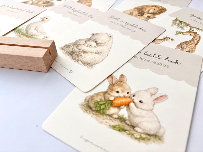 May include: A collection of illustrated cards featuring various animals, including a bear with a cub, a lion with a cub, and two rabbits sharing a carrot. The cards have scalloped edges and German text. A wooden card holder is in the foreground.
