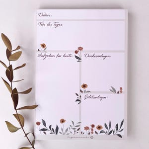 Könnte beinhalten: Ein weißer Notizblock mit Abschnitten für die Tagesplanung, verziert mit braunen und grünen Blumenillustrationen. Der Notizblock enthält handschriftlichen deutschen Text mit Abschnitten für Datum, Aufgaben und Dankbarkeit.