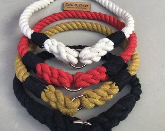May include: Four different colors of rope dog collars, white, red, gold, and black, with silver D-rings. The collars are stacked on top of each other. The top collar has a small wooden tag that says "DOG & Coast, Hand Crafted Dog Designs".