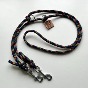 Pode incluir: Uma trela multicolorida para cães com um padrão de arco-íris. A trela tem dois fechos de metal prateado e uma pequena etiqueta de madeira que diz "Dog & Coast". A trela é feita de um material de corda durável.