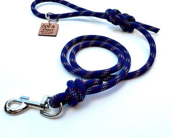 May include: A blue and red patterned dog leash with a silver metal clasp. The leash has a loop handle and a wooden tag that reads "DOG & Coast". The leash is made of a thick, durable rope material.