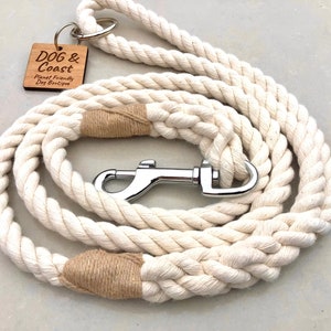 May include: A white cotton rope dog lead with a brown leather handle and a silver metal clip. The lead is coiled up and has a wooden tag attached to it that says "DOG & Coast Planet Friendly Dog Boutique".