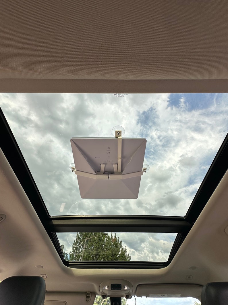 Peut inclure: Une tablette blanche fix&eacute;e au toit ouvrant d'une voiture &agrave; l'aide de ventouses. La tablette est inclin&eacute;e vers l'avant de la voiture. Le toit ouvrant est ouvert, r&eacute;v&eacute;lant un ciel bleu avec des nuages blancs.