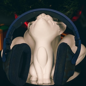May include: A white ceramic cat figurine with a blue and black headset resting on it. The headset has large, cushioned earcups. A wrapped gift box with a green bow is in the background.