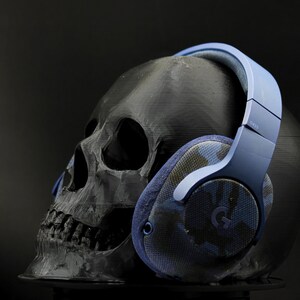 Puede incluir: Auriculares azules con estampado de camuflaje y diadema azul. Los auriculares descansan sobre una calavera negra.