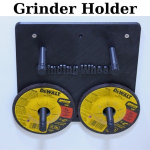 May include: A black wall-mounted grinder holder with two slots for holding grinding wheels. The holder has two black handles and the text "Grinding Wheel" is printed on the front. Two Dewalt grinding wheels are in the slots.