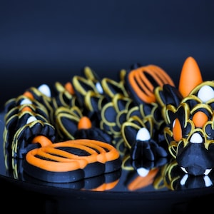 May include: A black and orange dragon sculpture with gold accents. The dragon has a detailed design with a stylized head, body, and tail. The sculpture is displayed on a reflective surface against a dark background.