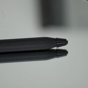 May include: A black stylus pen with a conical tip and a separate black stylus tip replacement. The pen is lying on a reflective surface, with its reflection visible. The replacement tip is a stack of black rings.