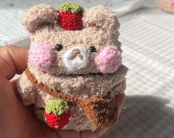 May include: A plush brown bear with pink cheeks and a white nose, wearing a green and red strawberry hat and a brown strap. The bear is holding a green and red strawberry in its paw.