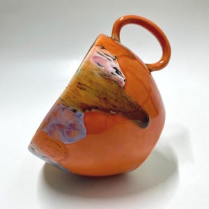Pode incluir: Uma caneca de cerâmica laranja vibrante com um design angular único. A caneca apresenta uma base arredondada e uma alça grande e circular. A parte superior tem um padrão abstrato colorido com tons de amarelo, rosa e azul, adicionando um toque artístico.