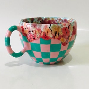 Może przedstawiać: Ceramiczny kubek w kratkę w kolorze różowym i turkusowym. Górna krawędź kubka jest ozdobiona kwiatowym wzorem w odcieniach różowego, czerwonego i żółtego.