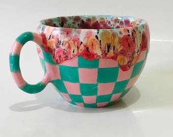 May include: A ceramic mug with a pink and teal checkered pattern. The top rim of the mug is decorated with a floral design in shades of pink, red, and yellow.