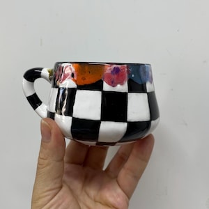 May include: A ceramic mug with a black and white chequered pattern on the body. The rim has a colourful, abstract design with orange, pink, and blue hues. The handle is also black and white chequered.
