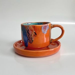 May include: A ceramic cup and saucer set featuring a vibrant orange glaze with abstract splashes of blue, purple, and white. The cup has a rounded body and a curved handle, while the matching saucer has a smooth, circular rim and a glossy finish.