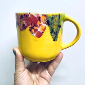 May include: A bright yellow ceramic mug with a rounded handle. The top edge features a colorful, textured glaze in shades of red, orange, and green. The mug is held against a white background.