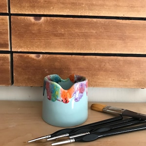Pode incluir: Um pequeno recipiente de cerâmica azul claro com uma colorida cobertura gotejante. Vários pincéis com cabos pretos e ponteiras prateadas estão dispostos nas proximidades. O recipiente está sobre uma superfície de madeira clara, com um fundo de painéis de madeira.
