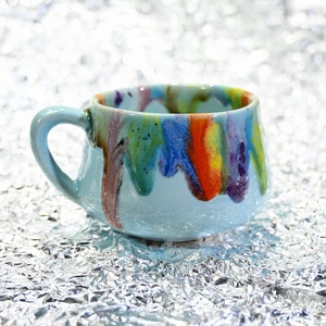 May include: A light blue ceramic mug with a rounded shape and a handle. The rim and interior are decorated with colorful drips of paint in shades of red, orange, yellow, blue, and purple. The mug sits on a textured silver surface.
