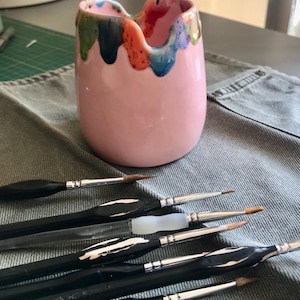 May include: A pink ceramic paint brush holder with colorful dripped paint around the rim. Several black-handled paint brushes with silver ferrules and fine tips are arranged in front of the holder, suggesting an art studio setting.