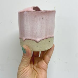 Könnte beinhalten: Eine handgefertigte Keramik-Tasse mit einem einzigartigen Design. Die obere Hälfte ist ein zartes Rosa mit einem tropfenden Glanzeffekt, während die untere Hälfte ein natürliches, gesprenkeltes Beige ist. Die Tasse hat eine gebogene Öffnung.