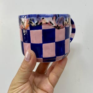 Może przedstawiać: Ceramiczny kubek w różowo-granatową kratę. Obręcz zdobi teksturowany, kwiatowy wzór z różowymi, niebieskimi i pomarańczowymi akcentami. Kubek ma zakrzywiony uchwyt i jest trzymany przez dłoń.