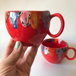 Op de afbeelding: Twee rode keramische mokken met kleurrijk druipglazuur rond de randen. De grotere mok wordt in een hand gehouden en de kleinere mok staat ernaast. De glazuurkleuren zijn onder meer blauw, oranje, geel en zwart.