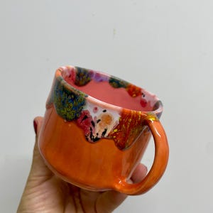 Pode incluir: Caneca de cerâmica laranja com uma alça curva. A borda apresenta uma cobertura colorida escorrida em tons de azul, verde, vermelho e amarelo. O interior da caneca é rosa claro.