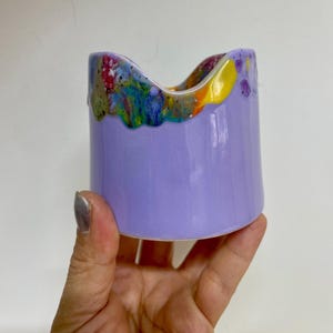 May include: A handcrafted ceramic container with a light purple exterior and a colorful, abstract rim. The rim features a mix of colors including red, yellow, blue, and green. The container is cylindrical with a slightly curved top.