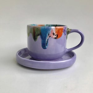 Puede incluir: Un juego de taza y platillo de cerámica púrpura con un diseño de glaseado de goteo colorido. El glaseado es una mezcla de azul, verde, naranja y rosa.