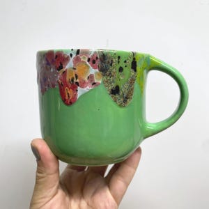 May include: A handmade ceramic mug with a vibrant green exterior and a colorful, dripped glaze around the rim. The glaze features red, yellow, and black accents. The mug has a rounded handle.