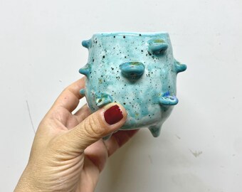 May include: A small, turquoise ceramic pot with a speckled finish and raised, rounded protrusions. The pot has three small feet and is held against a white background. The pot's color is a vibrant, speckled turquoise.