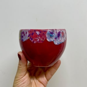 May include: A vibrant, red ceramic bowl with a distinctive, textured rim showcasing a mix of purple, pink, and blue shades. The bowl is held in a hand against a plain white backdrop. This decorative bowl is suitable for serving or display.