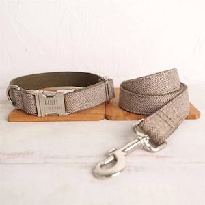 Op de afbeelding: Een bruine en groene hondenhalsband en riem in herringbone-patroon met een zilveren gesp en een gepersonaliseerd label met de tekst "BAILEY 123-456-1890".