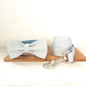 Op de afbeelding: Een lichtblauwe en witte gingham hondenhalsband en riemset met een bijpassende strik. De halsband en riem zijn omkeerbaar met een effen lichtblauwe zijde.