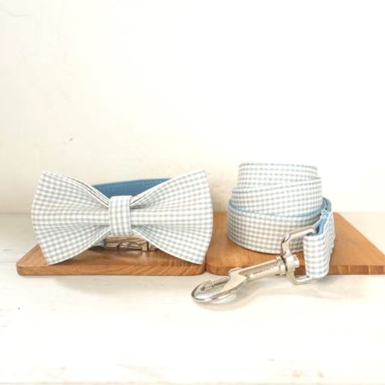 Op de afbeelding: Een lichtblauwe en witte gingham hondenhalsband en riemset met een bijpassende strik. De halsband en riem zijn omkeerbaar met een effen lichtblauwe zijde.