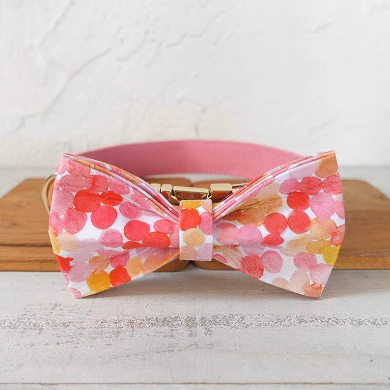May include: A pink dog collar with a bow tie featuring a watercolor pattern of pink, orange, and yellow circles on a white background.