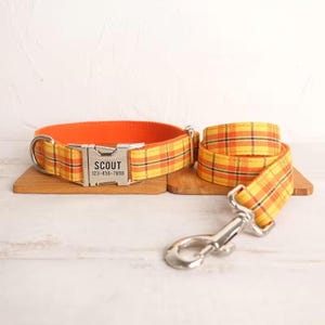 May include: Orange and yellow plaid dog collar and leash set with a silver buckle and a personalized tag that reads "SCOUT 123-456-7890".